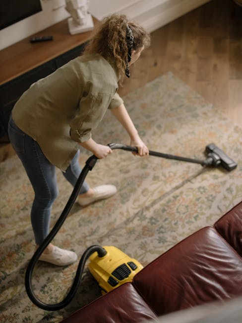 A woman with curly hair, wearing a light-colored jacket, dark jeans, and white sneakers, is performing surface cleaning using a yellow vacuum cleaner with a hose attachment, on a patterned area rug in a residential living room. The room features wooden flooring and a leather sofa, with natural lighting illuminating the space. The scene depicts thorough deep cleaning and sanitisation, highlighting the use of professional tools by Deep Cleaning Enfield to maintain hygiene in domestic environments, consistent with the service offered near Enfield Chase station.
