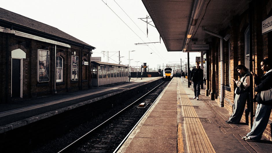 An image of a train station platform featuring a brick building on the right side with large windows, where several passengers are waiting, some standing and others leaning against the wall. The platform surface appears clean and dry, with a tactile paving strip along the edge near the railway tracks. On the opposite platform, a modern train with yellow and white colors is approaching, with overhead electric wires visible above the tracks. The station is illuminated by natural daylight, creating a well-lit environment that highlights the tidy, well-maintained surfaces typical of professional cleaning standards. Deep Cleaning Enfield specializes in detailed surface cleaning and sanitisation, ensuring that areas such as train stations and other public spaces are hygienic and safe for visitors.