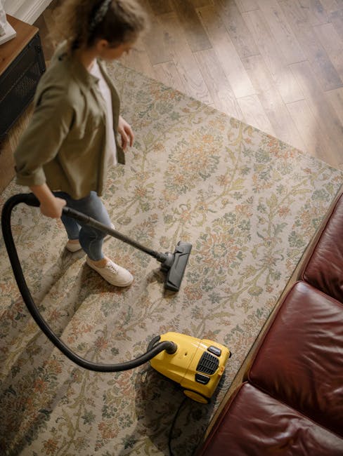 A woman with curly hair, wearing a light-colored jacket, dark jeans, and white sneakers, is performing surface cleaning using a yellow vacuum cleaner with a hose attachment, on a patterned area rug in a residential living room. The room features wooden flooring and a leather sofa, with natural lighting illuminating the space. The scene depicts thorough deep cleaning and sanitisation, highlighting the use of professional tools by Deep Cleaning Enfield to maintain hygiene in domestic environments, consistent with the service offered near Enfield Chase station.
