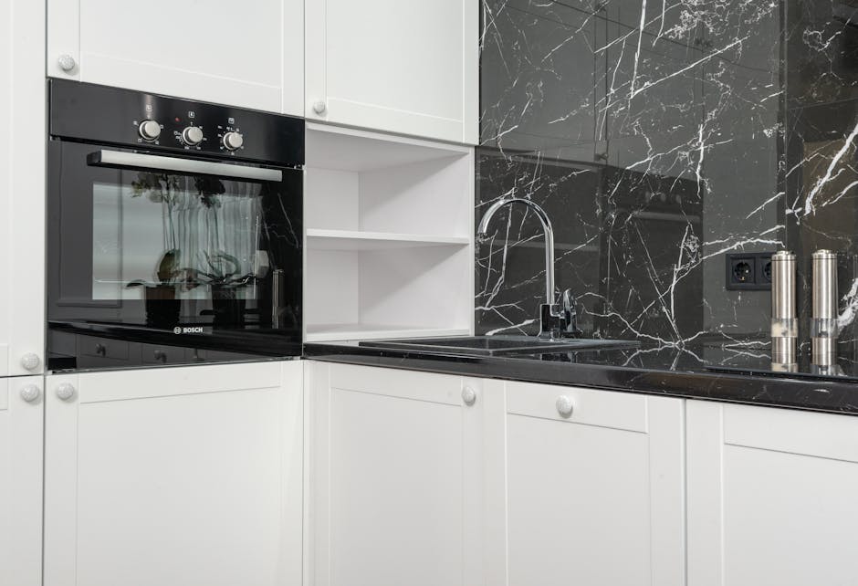 A modern kitchen surface featuring a black marble countertop with white veining, adjacent to a stainless steel sink with a chrome faucet. The black marble backsplash extends behind the sink and countertop, providing a sleek and polished appearance. To the left, a built-in oven with a black glass door and control knobs is integrated into white cabinetry with round white handles, some airily open shelving is visible above the countertop. The workspace appears clean and well-maintained, showcasing the effectiveness of surface cleaning and sanitisation practices by Deep Cleaning Enfield within a bright room illuminated by natural light or overhead fixtures.