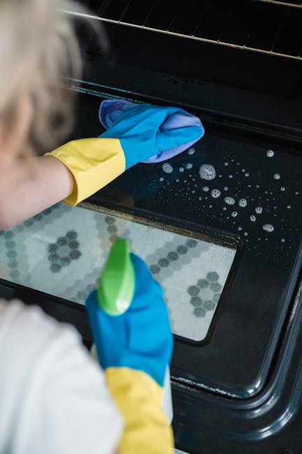 A person wearing blue and yellow rubber cleaning gloves is cleaning the glass surface of a black oven door with soapy water and a cloth. The oven's interior, visible through the glass, shows a clean, reflective surface with some bubbles and foam present. The setting is a domestic kitchen environment, focusing on surface cleaning and maintenance as part of deep cleaning processes. Deep Cleaning Enfield provides professional domestic cleaning services, including oven sanitisation and surface cleaning, to ensure hygiene and thorough cleanliness in busy households.