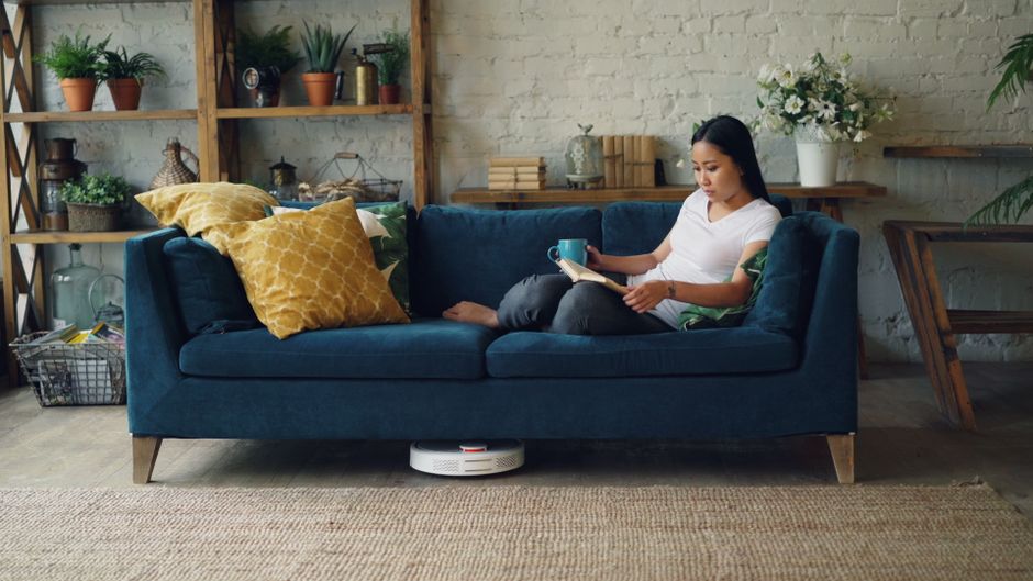 A person in a light-colored sleeveless top is using their hands to straighten a large beige cushion on a light-colored sofa in a living room. The sofa has matching cushions and is positioned against a